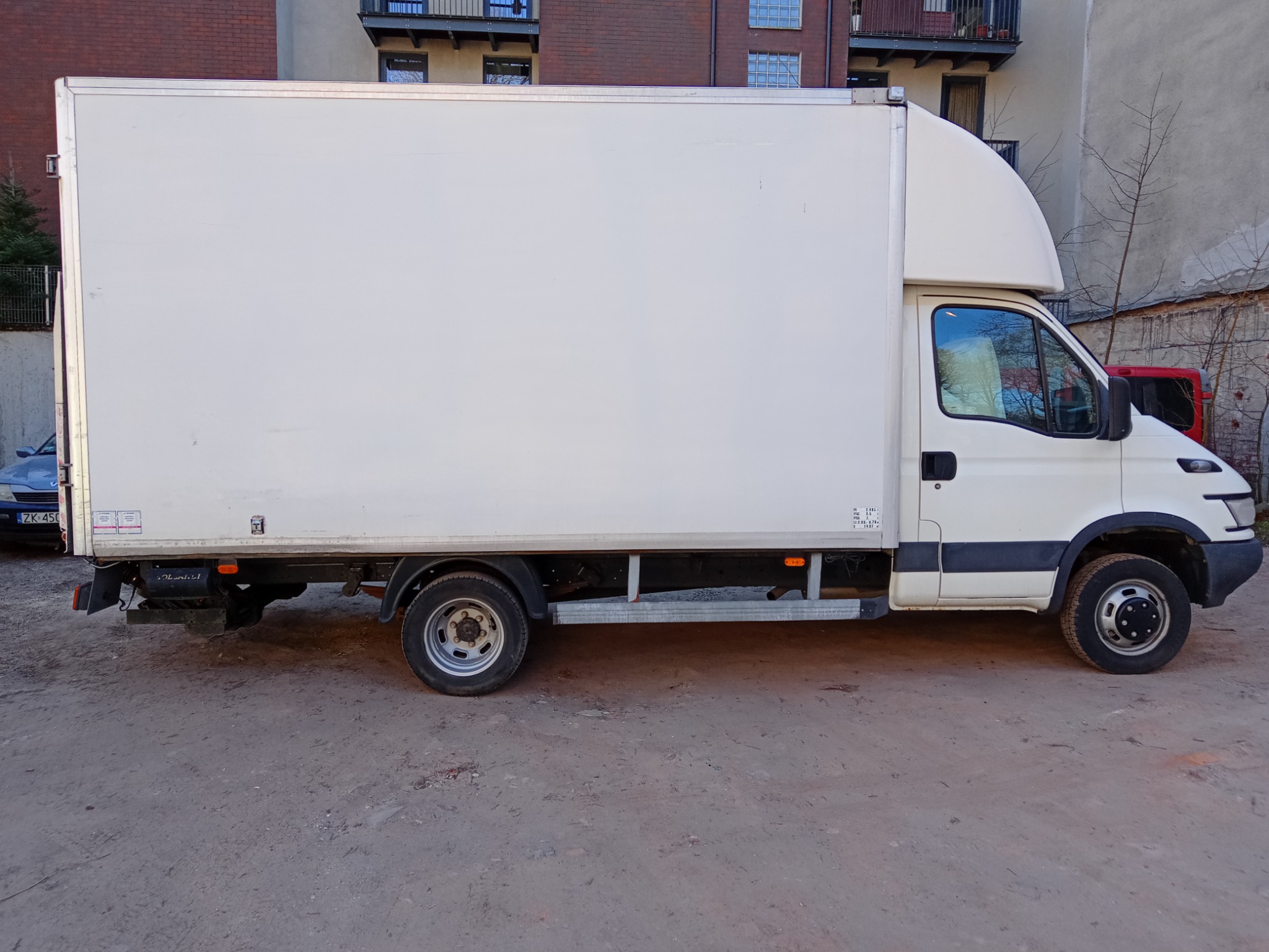 Biały samochód dostawczy z zabudową kontenerową zaparkowany na placu, widok z boku, w tle budynki mieszkalne.