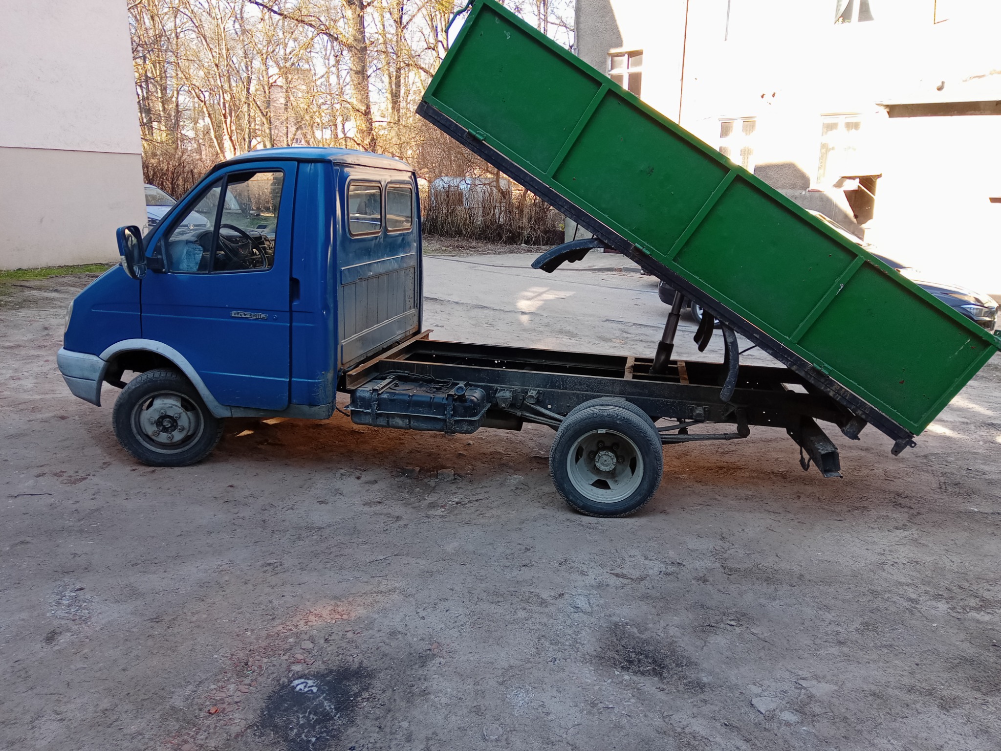 Niebieski samochód dostawczy z podniesioną, zieloną skrzynią ładunkową na podwórzu.