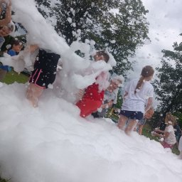 Bajlandia - Dzieci bawią się w pianie na trawie, w tle drzewa i niebo. Piana częściowo zasłania widok, tworząc wrażenie zanurzenia w niej. Radosna zabawa w plenerze.