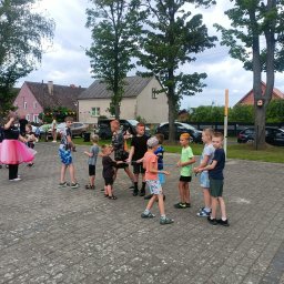 Bajlandia - Grupa dzieci gra w przeciąganie liny na zewnątrz, nadzorowana przez osobę w różowej tutu. Widoczne budynki i drzewa w tle, tworzące scenerię wiejskiego podwórka.