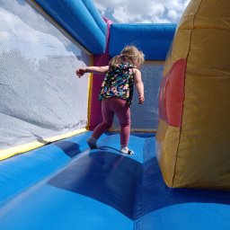 Bajlandia - Dziewczynka w kolorowej bluzce i różowych legginsach skacze w niebiesko-żółtym dmuchanym zamku z siatką zabezpieczającą. Słoneczny dzień, częściowo widoczne niebo.