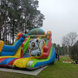 Bajlandia - Kolorowy dmuchany zamek ze zjeżdżalniami, z motywem zwierząt, ustawiony na trawie w otoczeniu drzew. Widoczne osoby i elementy infrastruktury.