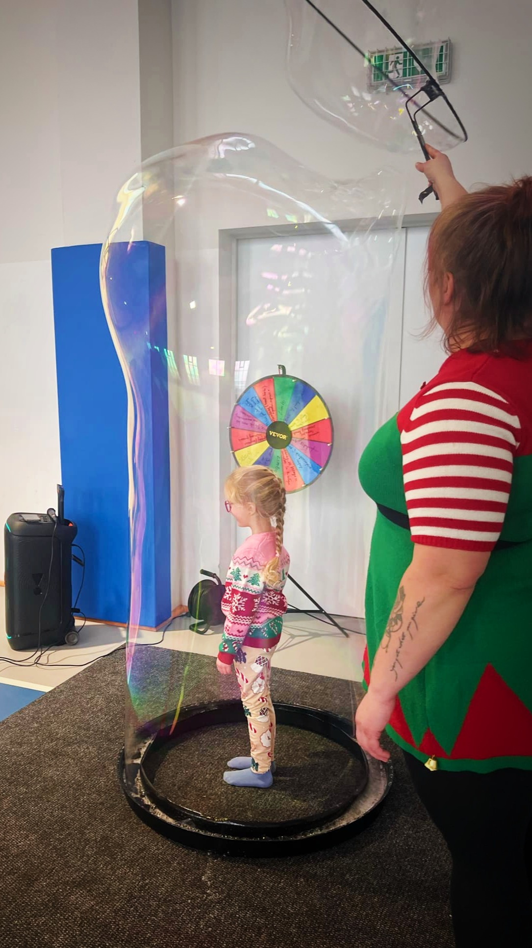 Dziewczynka w świątecznym swetrze i leginsach stoi wewnątrz ogromnej bańki mydlanej, tworzonej przez animatora w stroju elfa, w tle kolorowe koło fortuny i głośnik.