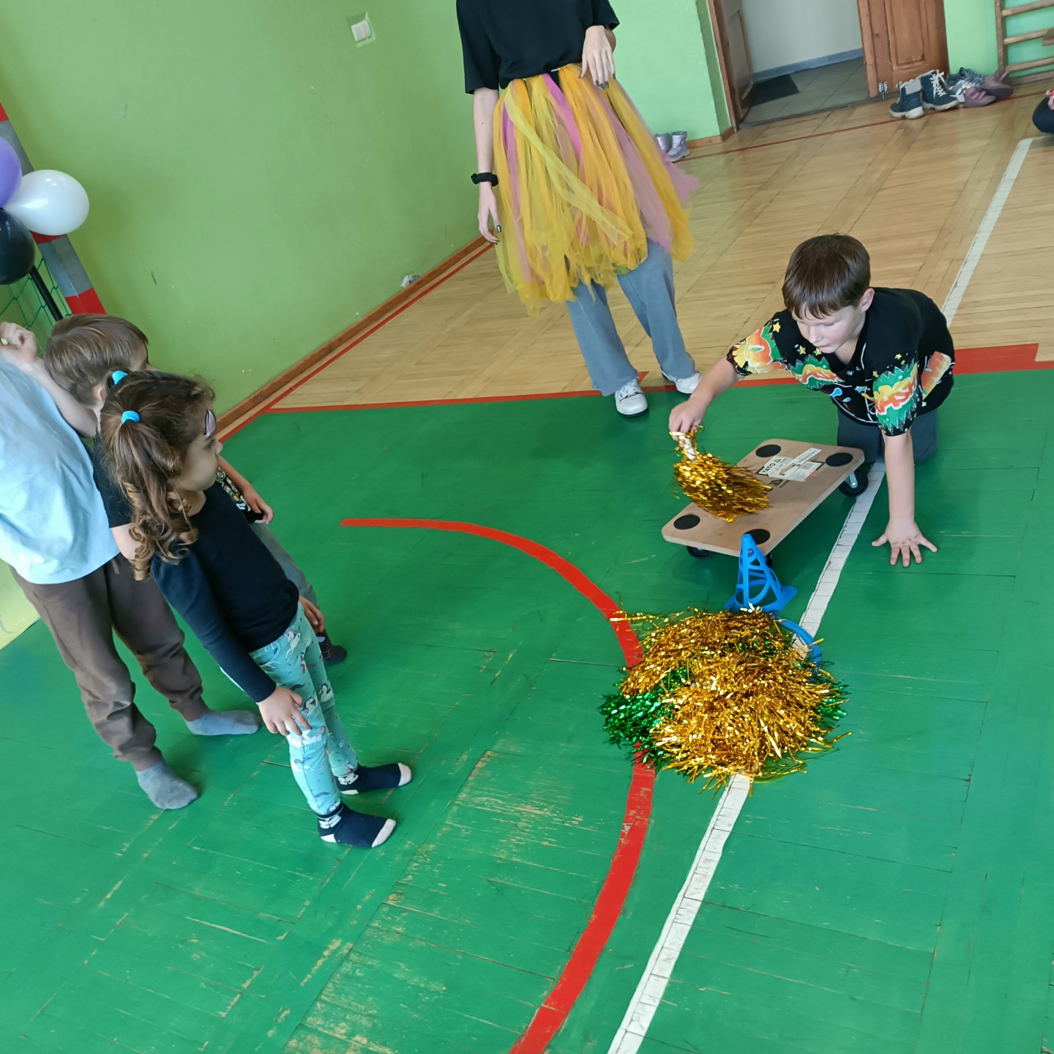 Dzieci uczestniczą w zabawie z pomponami i drewnianą platformą na kółkach na zielonej podłodze w hali sportowej, nadzorowane przez osobę w kolorowej spódnicy.