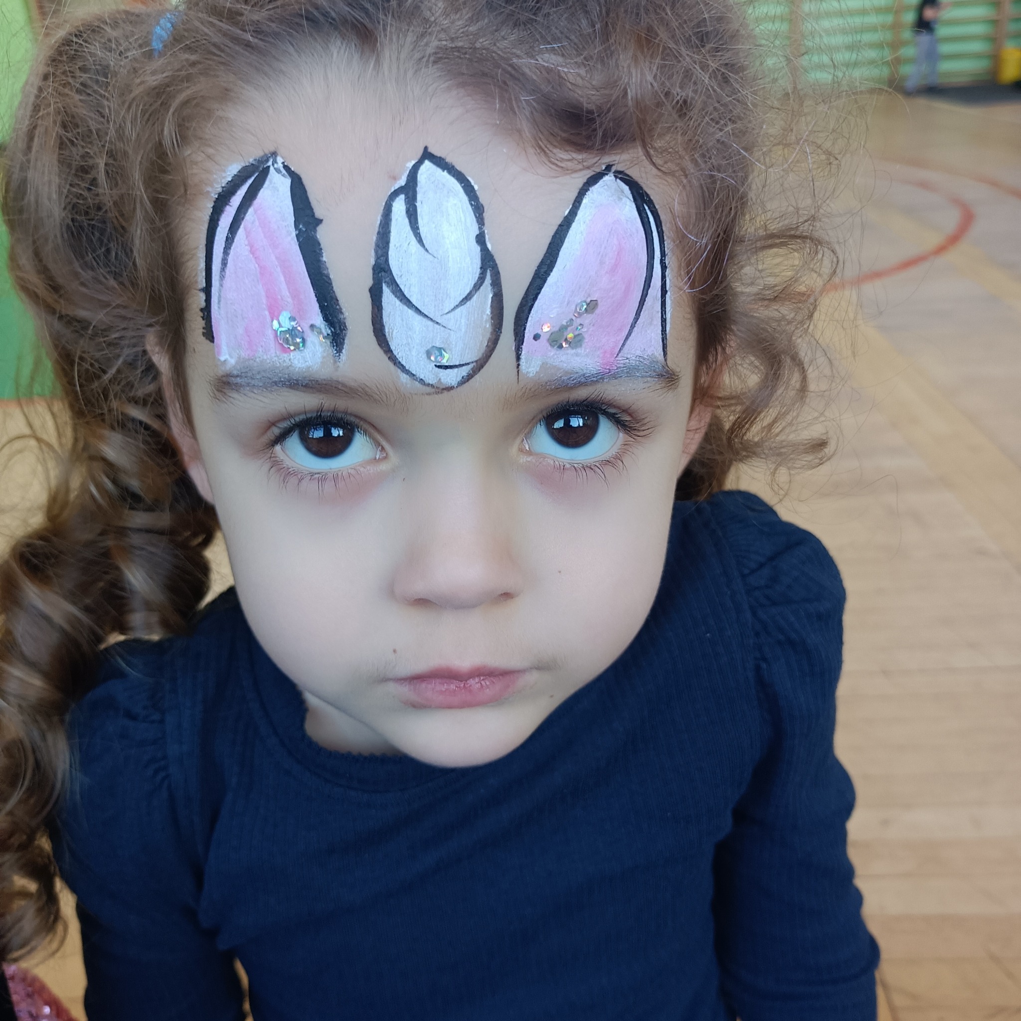 Dziewczynka z pomalowaną twarzą w uszy królika z brokatem na czole, patrząca w górę.