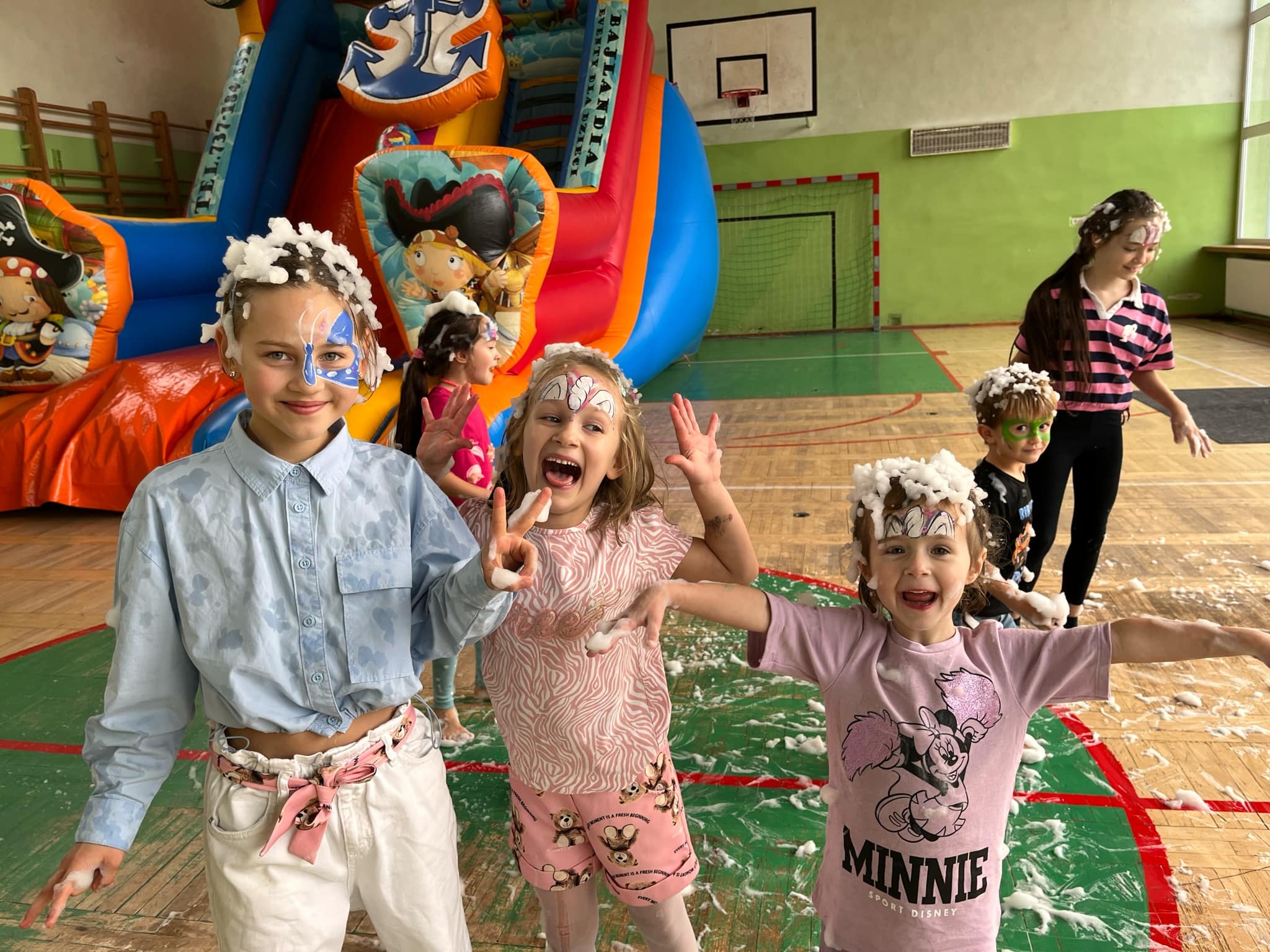Grupa roześmianych dzieci z pomalowanymi twarzami i pianą na włosach, pozuje na tle kolorowego dmuchańca w sali gimnastycznej.