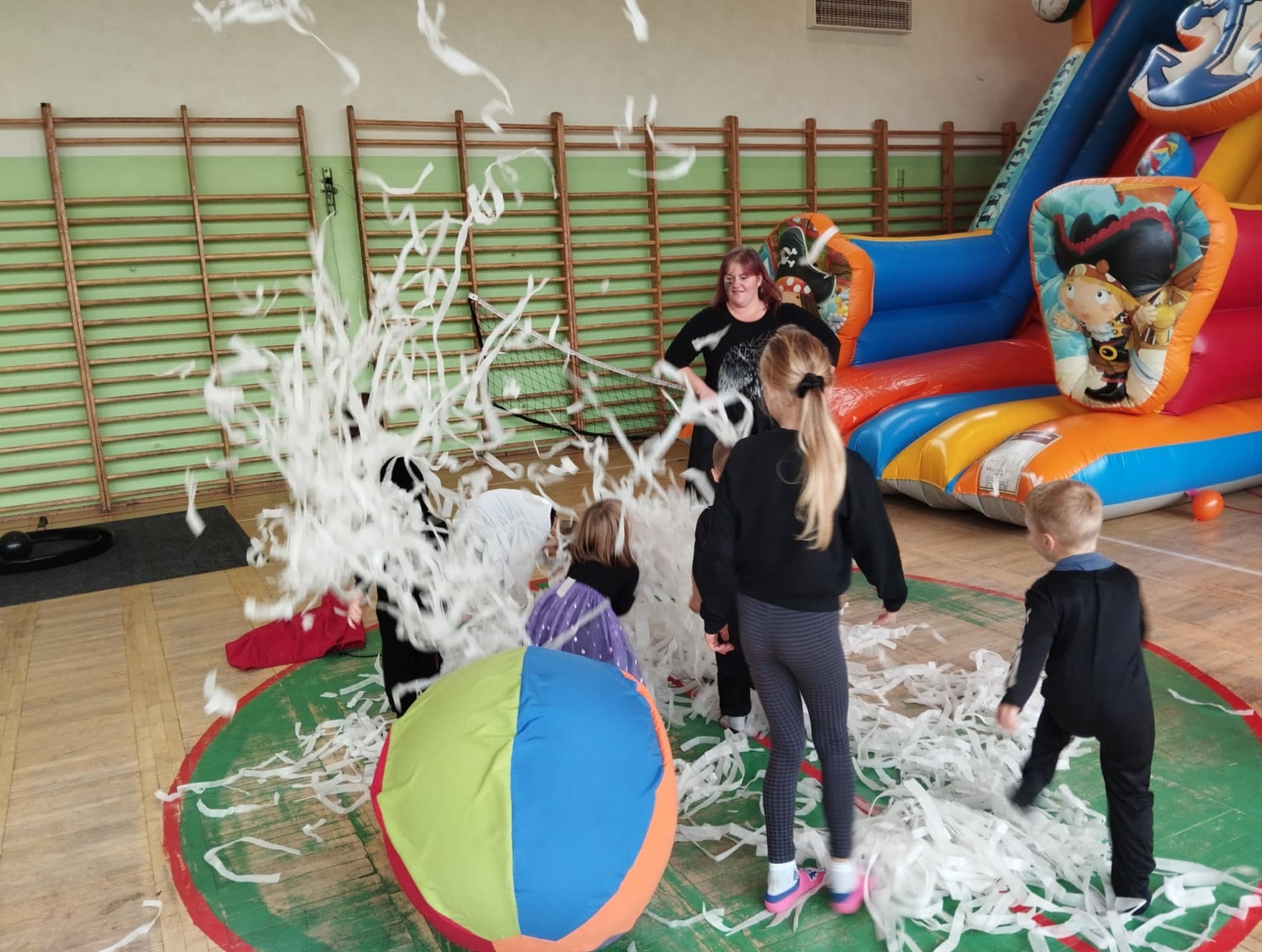 Dzieci bawią się w sali gimnastycznej z konfetti, w tle dmuchany zamek i drabinki gimnastyczne.