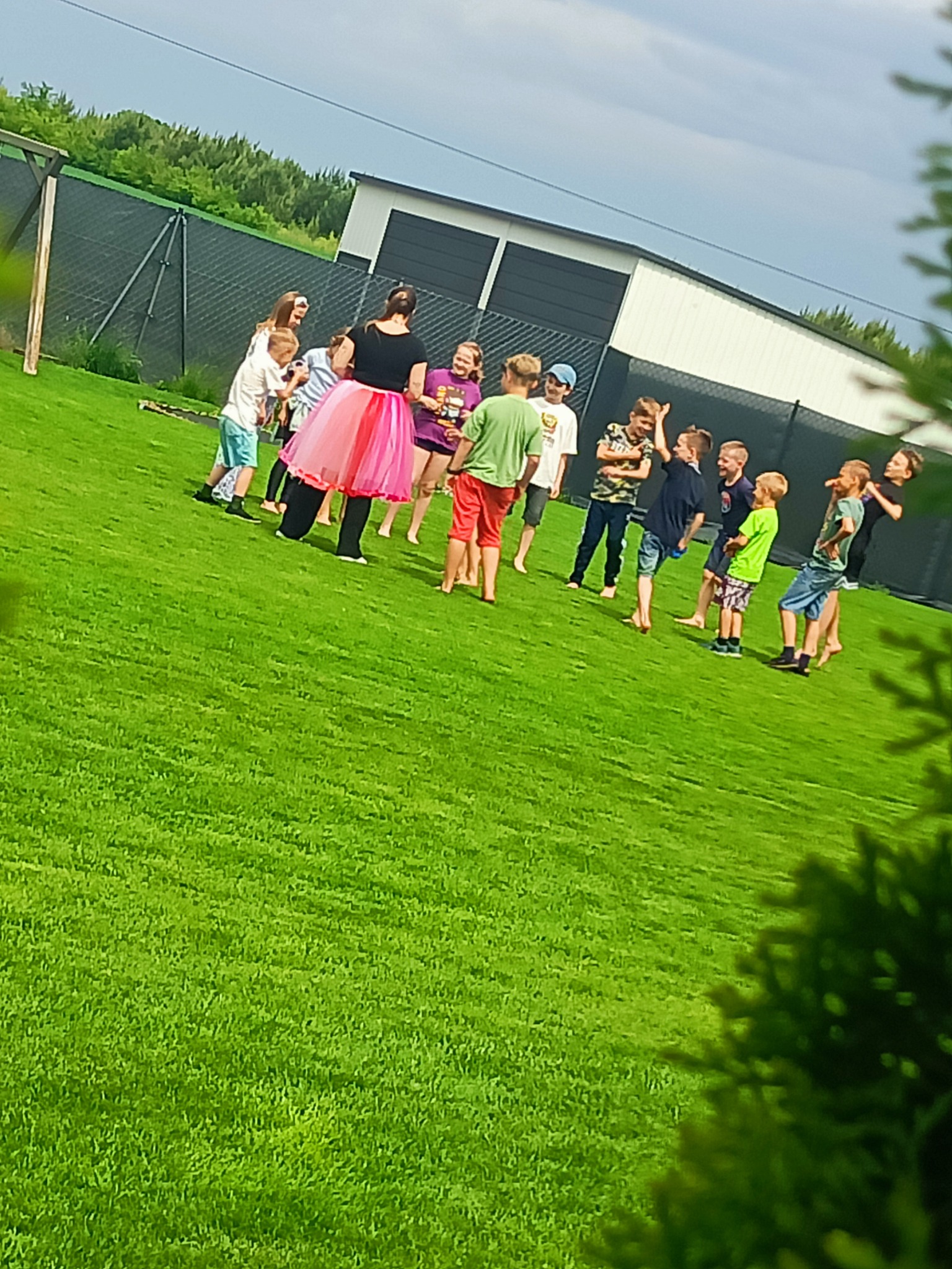 Animator w różowej tutu prowadzi zabawę na zielonej trawie z grupą dzieci podczas imprezy plenerowej. W tle widoczny budynek i ogrodzenie.