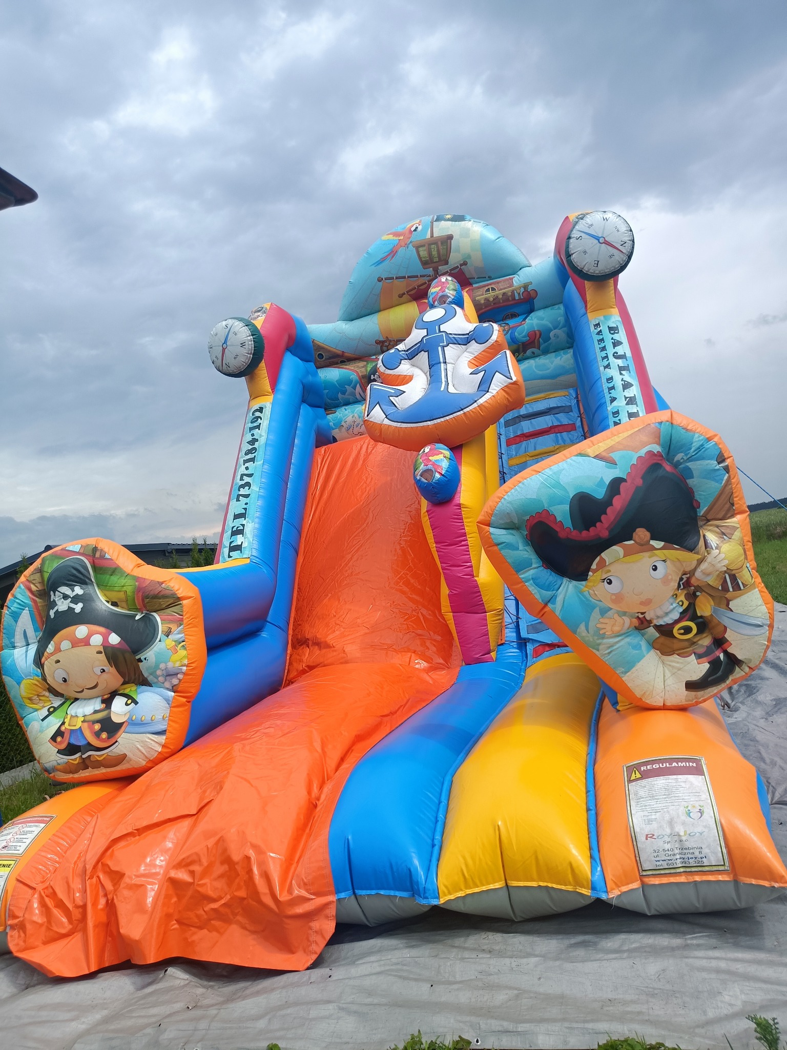 Kolorowa, dmuchana zjeżdżalnia w stylu pirackim z motywami kompasu, kotwicy i postaci piratów, ustawiona na trawie pod pochmurnym niebem.