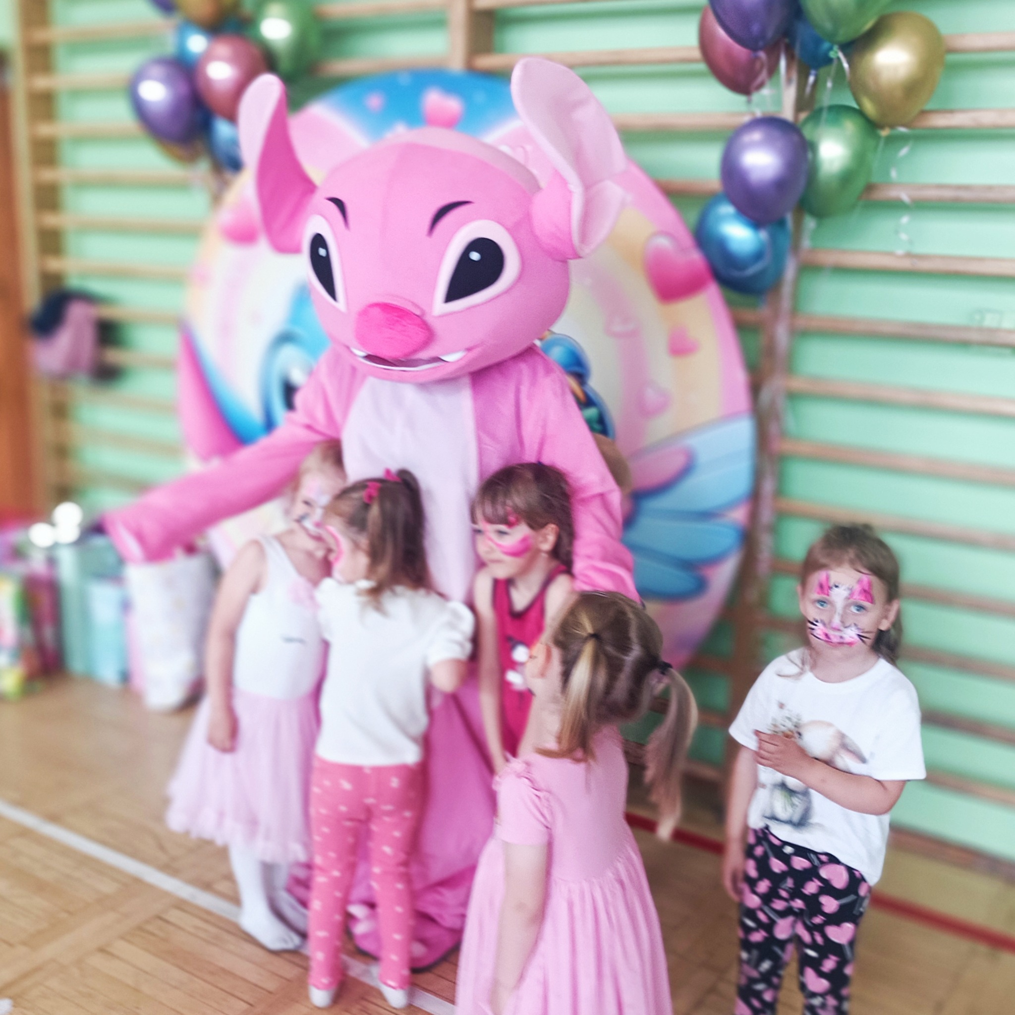 Animator w kostiumie różowego stwora z dziećmi z pomalowanymi twarzami na tle dekoracji balonowych i ściany drabinkowej w sali gimnastycznej.