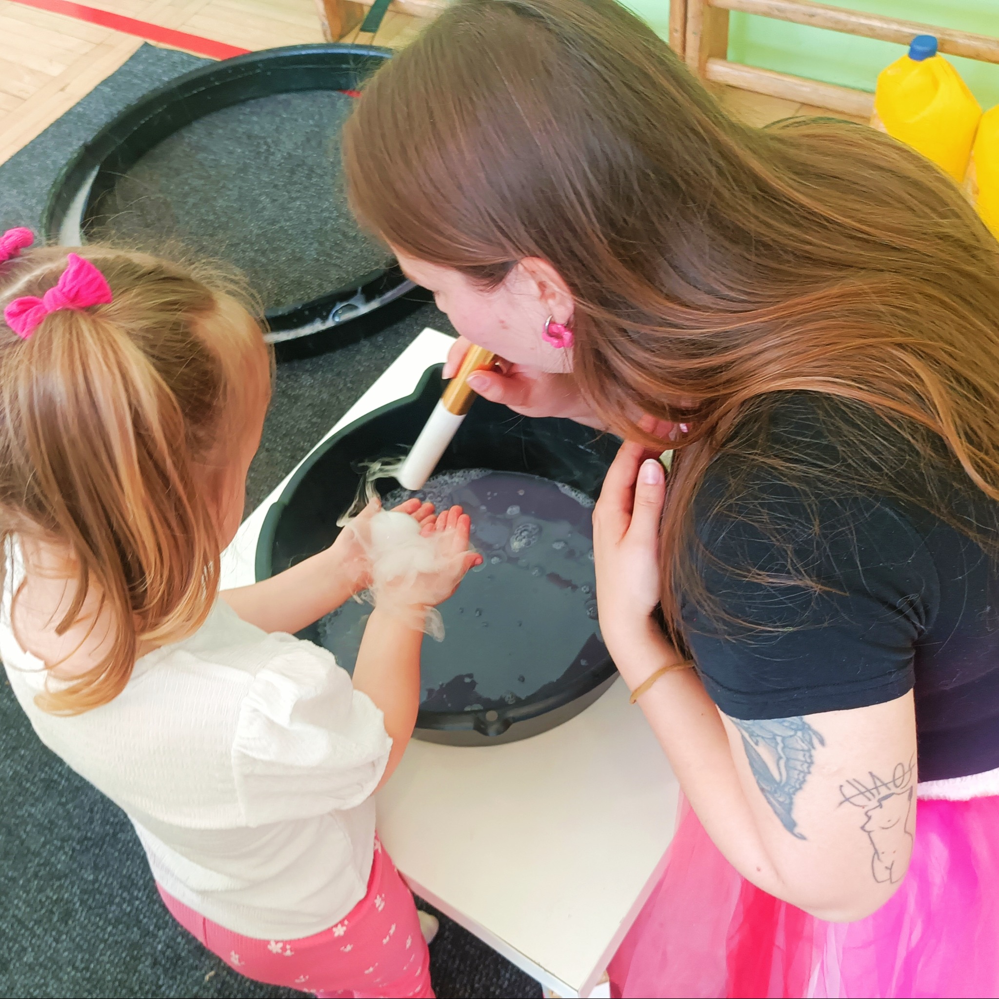 Animator dmucha bańki mydlane na dłonie dziewczynki w różowym stroju. Zabawa w pomieszczeniu z elementami drewnianymi i żółtymi pojemnikami w tle.