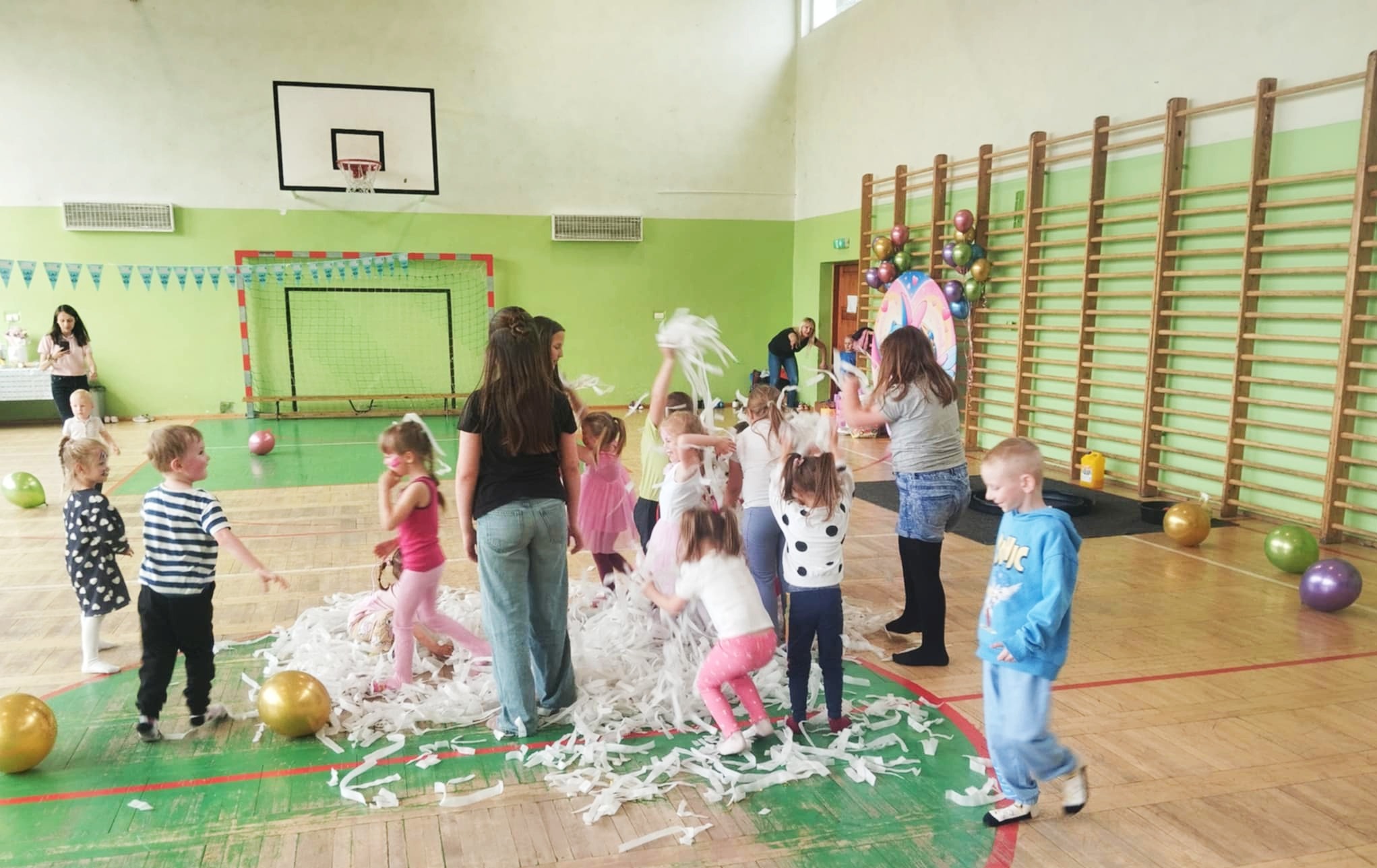 Grupka dzieci w sali gimnastycznej bawi się w konfetti; w tle drabinki gimnastyczne, kosz do koszykówki i dekoracje balonowe. Zabawa z animatorem.
