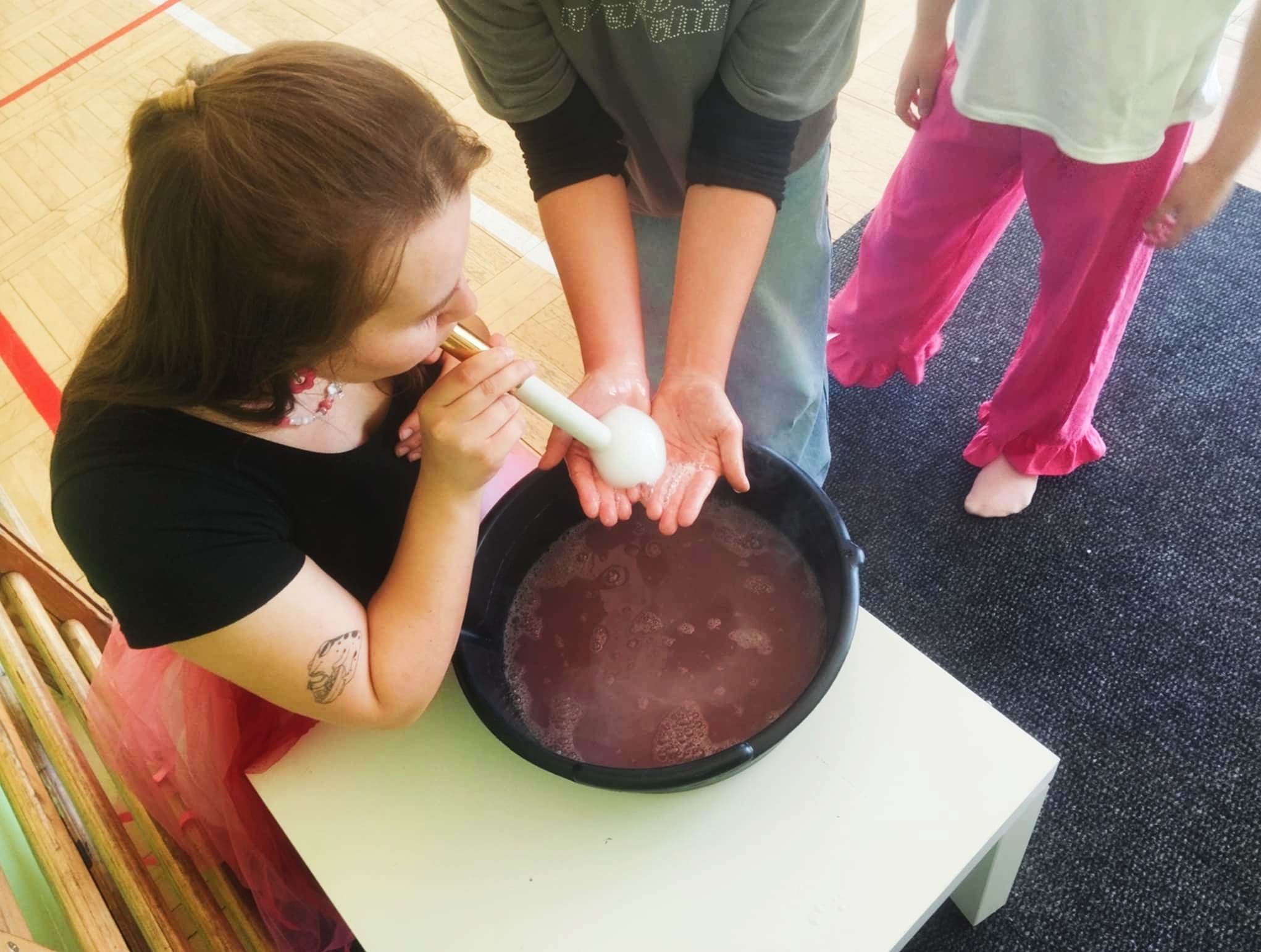 Kobieta z tatuażem na ramieniu dmucha w rurkę, tworząc dużą bańkę mydlaną nad miską. Dziecko z różowych spodniach obserwuje proces.