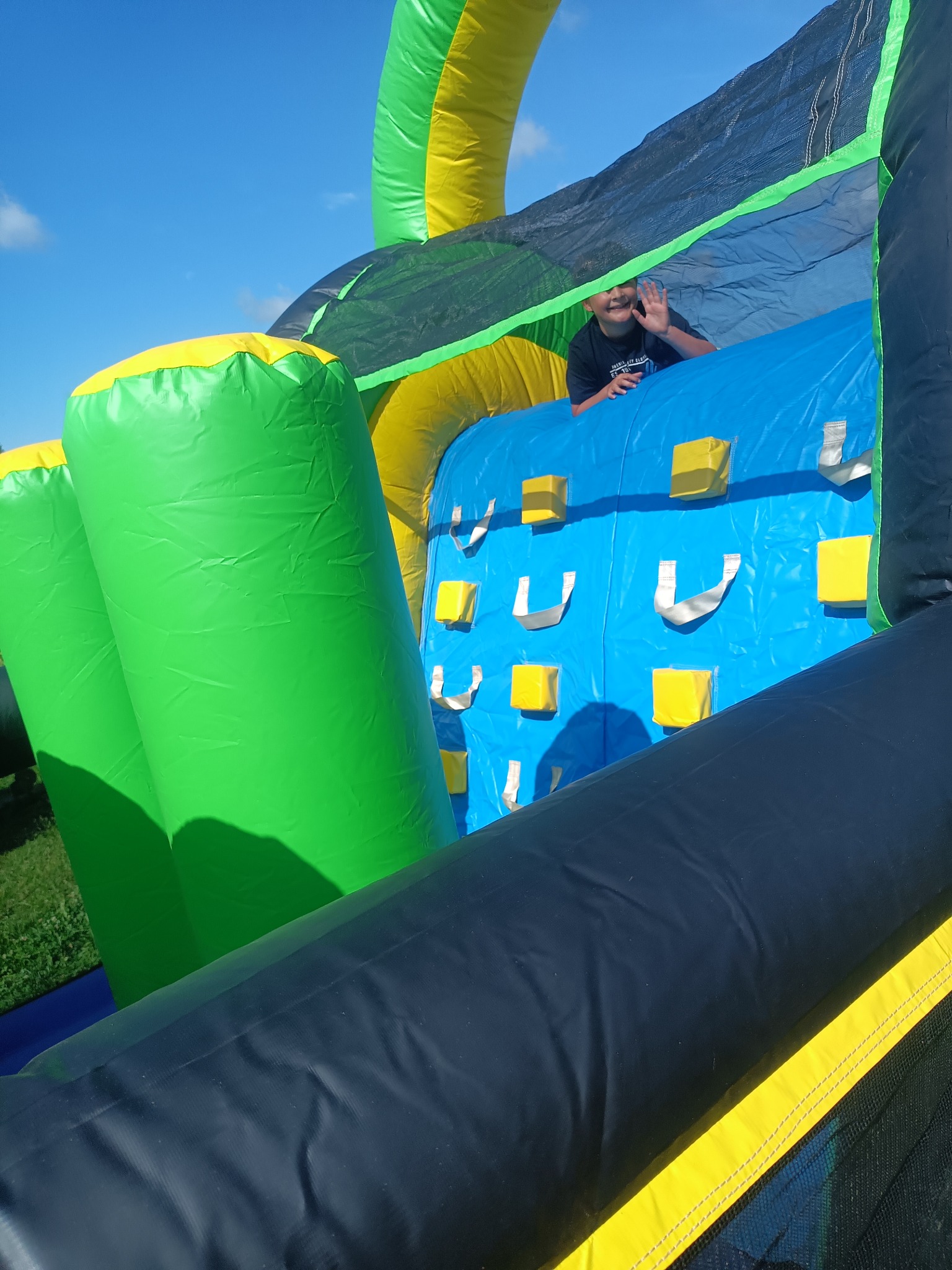 Chłopiec macha z dmuchanego zamku z elementami wspinaczki, w kolorach zielonym, żółtym, niebieskim i czarnym. Widok na błękitne niebo w tle.