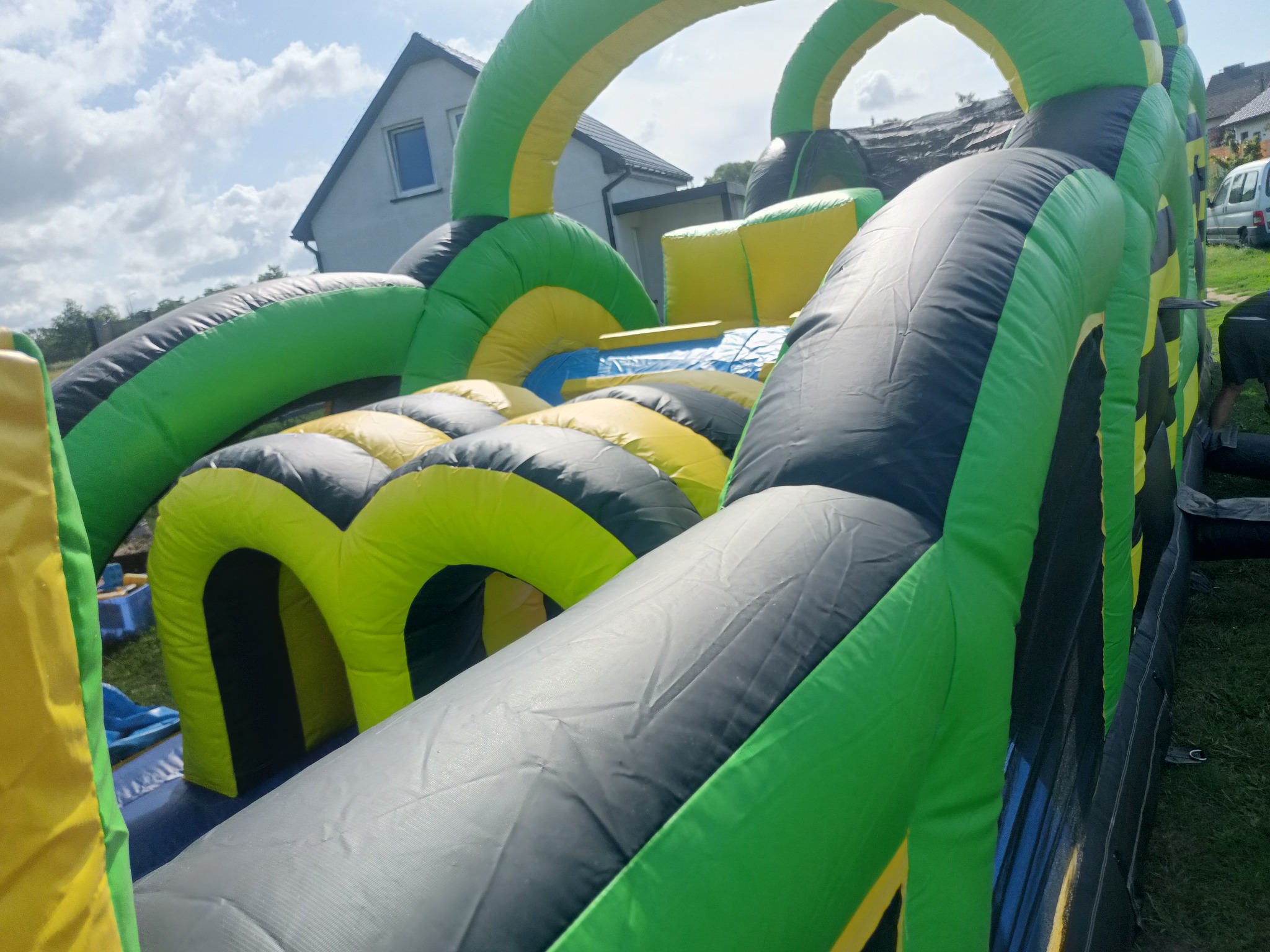 Kolorowy dmuchany tor przeszkód z zielono-żółto-szarymi elementami, ustawiony na trawie, z fragmentem domu w tle, widziany z perspektywy bliskiej ziemi.