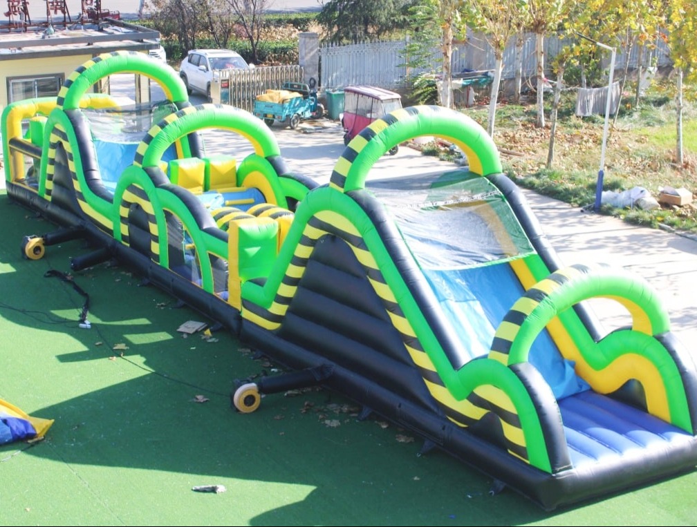 Długi, dmuchany tor przeszkód w kolorach zieleni, czerni i żółci, z elementami ślizgawki wodnej. Widoczne drzewa w tle.