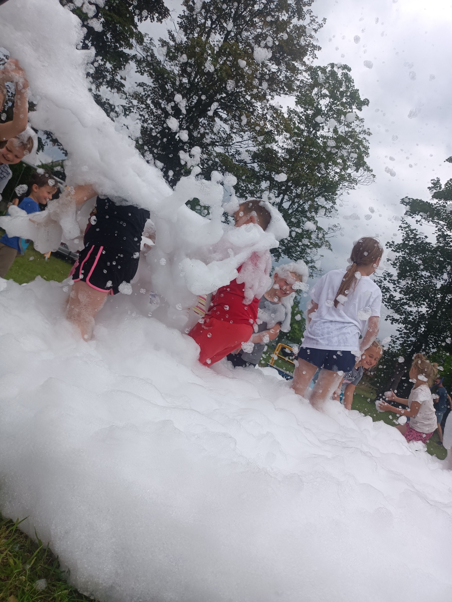 Dzieci bawią się w pianie na trawie, w tle drzewa i niebo. Piana częściowo zasłania widok, tworząc wrażenie zanurzenia w niej. Radosna zabawa w plenerze.