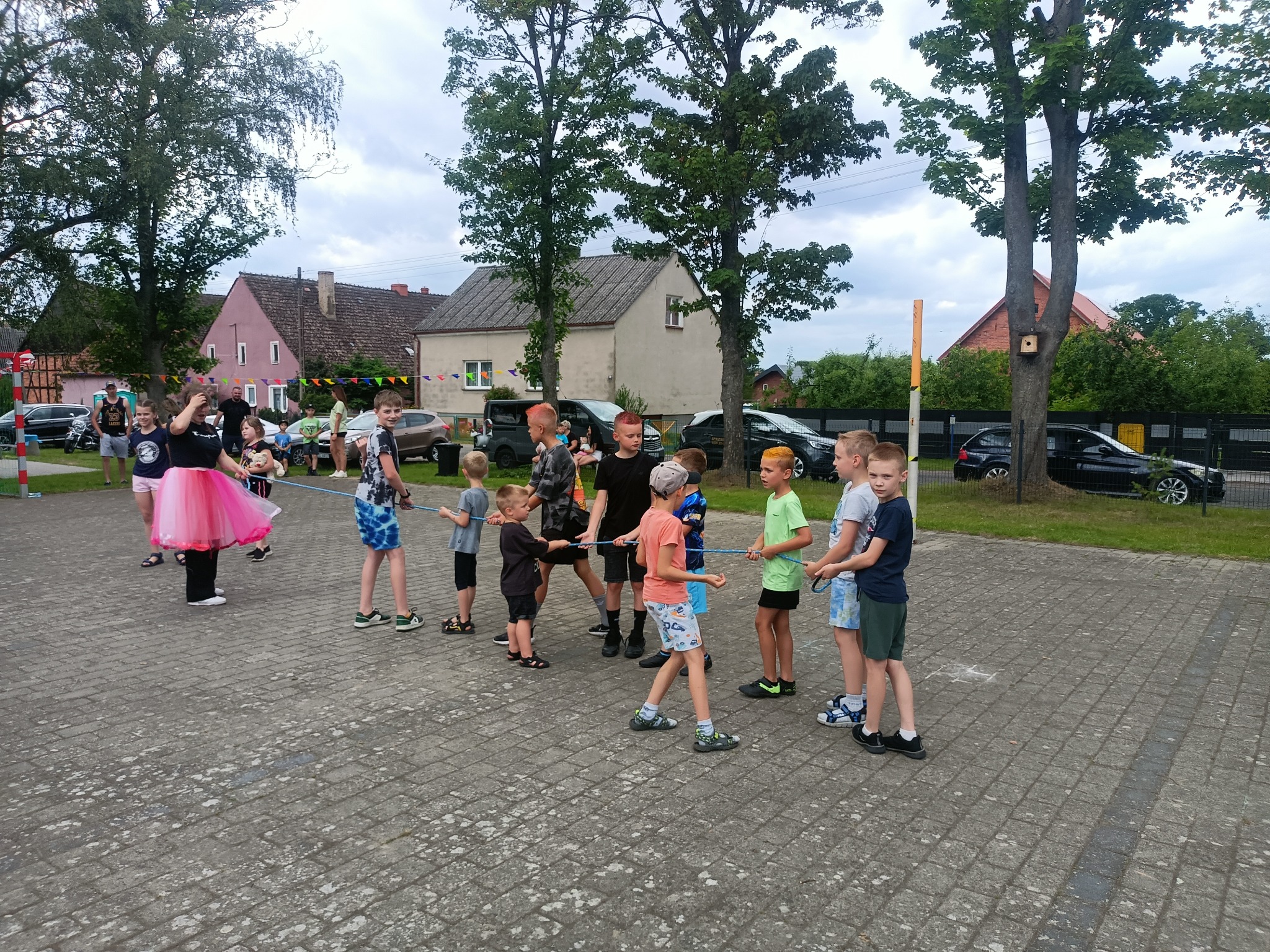 Grupa dzieci gra w przeciąganie liny na zewnątrz, nadzorowana przez osobę w różowej tutu. Widoczne budynki i drzewa w tle, tworzące scenerię wiejskiego podwórka.