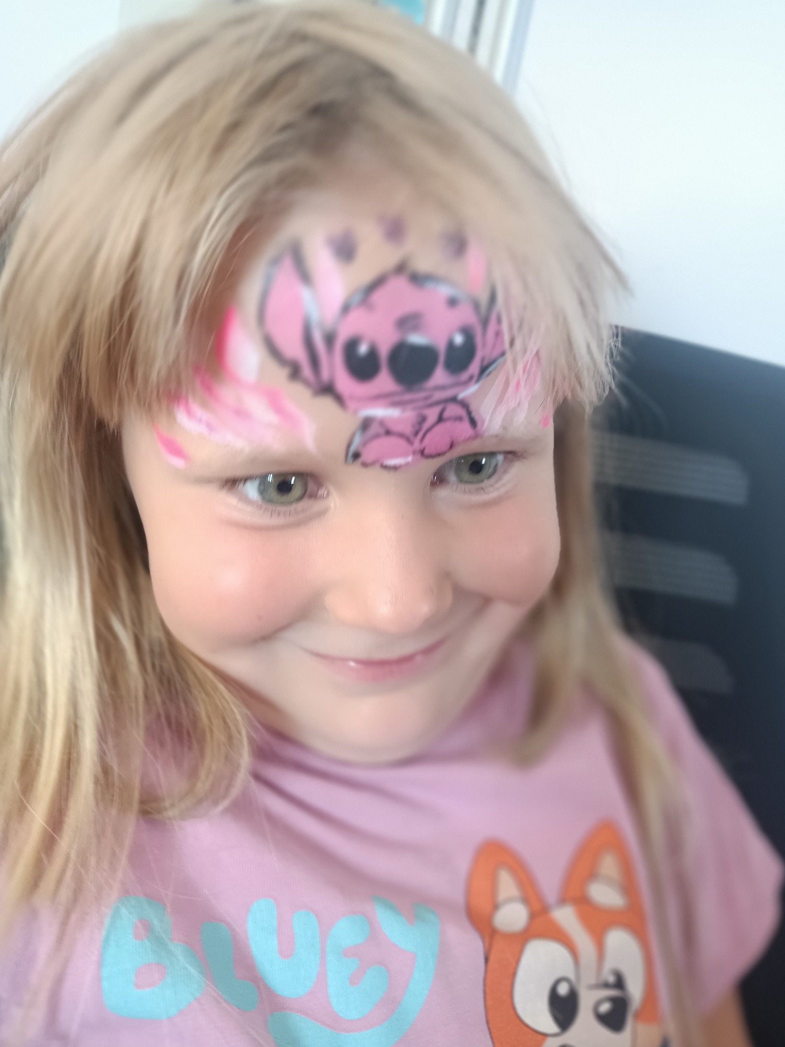 Dziewczynka z pomalowaną twarzą w różowego Sticha i różowe akcenty na czole, uśmiechnięta, w koszulce z postacią z bajki Bluey.