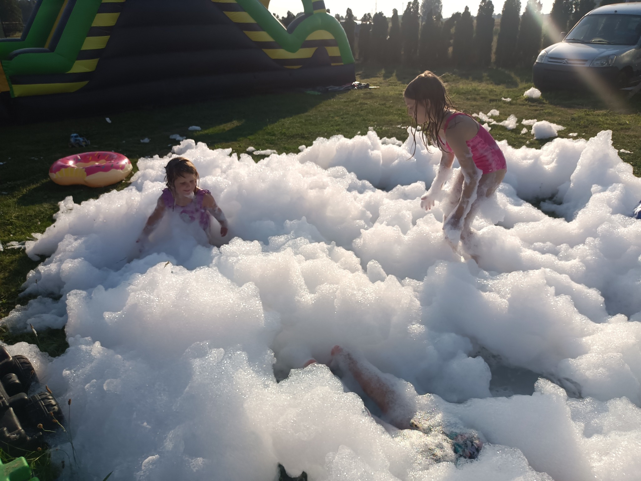 Dwie dziewczynki w różowych strojach bawią się w pianie na trawie, w tle dmuchaniec, auto i drzewa. Ujęcie z dołu, podkreślające dynamikę zabawy.