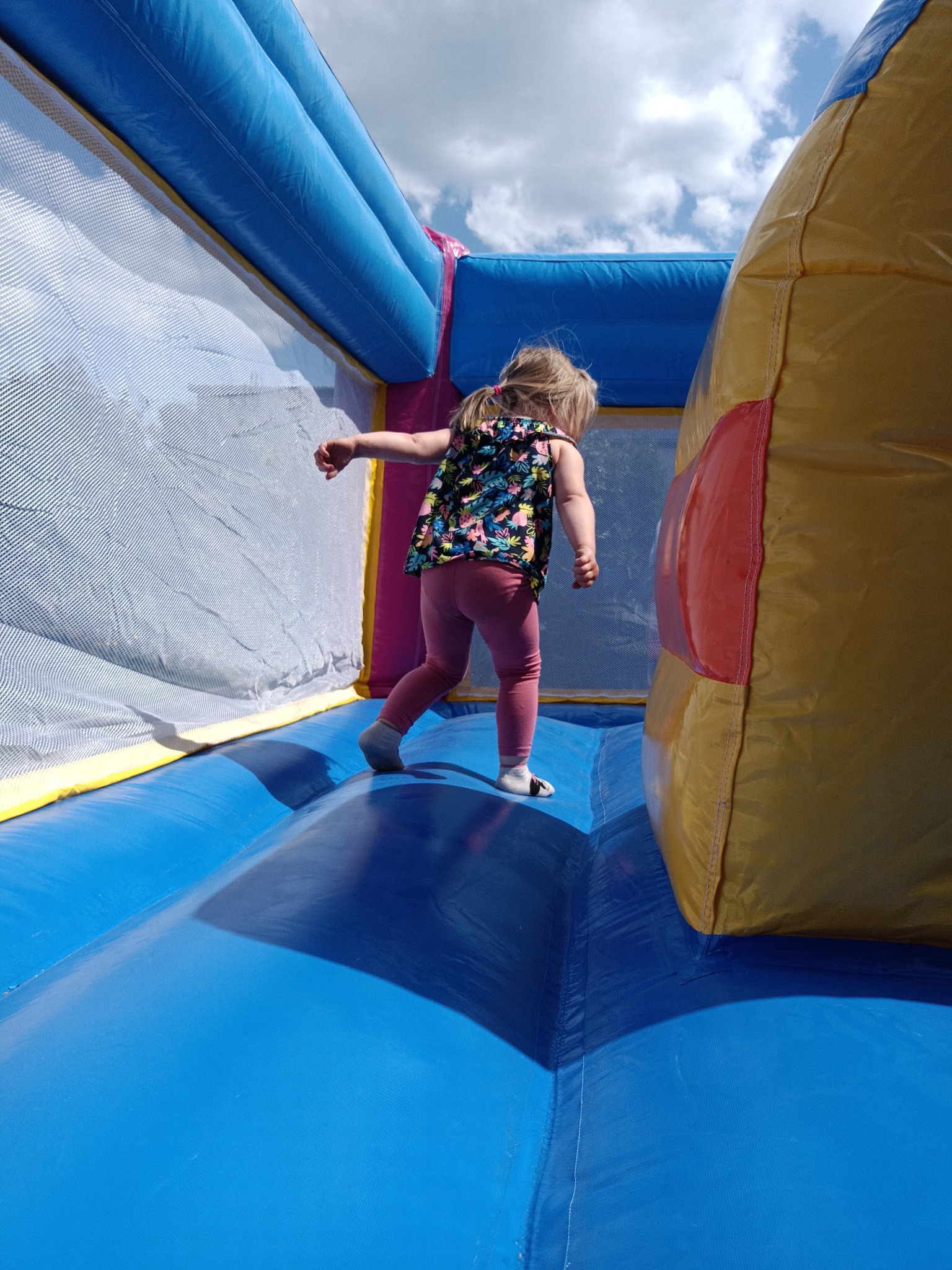 Dziewczynka w kolorowej bluzce i różowych legginsach skacze w niebiesko-żółtym dmuchanym zamku z siatką zabezpieczającą. Słoneczny dzień, częściowo widoczne niebo.
