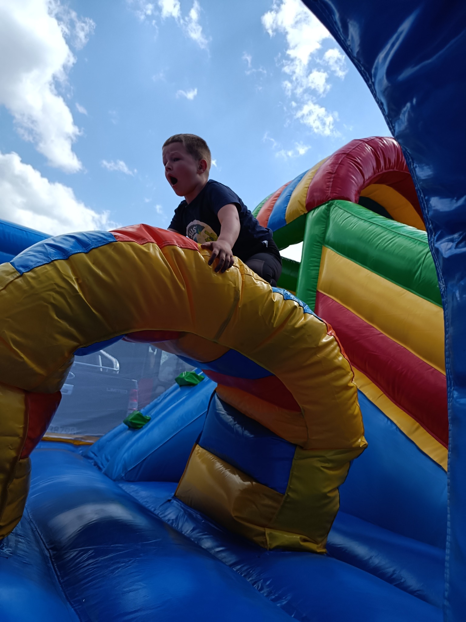 Chłopiec z otwartymi ustami wspina się na kolorowy dmuchany zamek na tle nieba z chmurami. Widok z dołu, intensywne barwy.