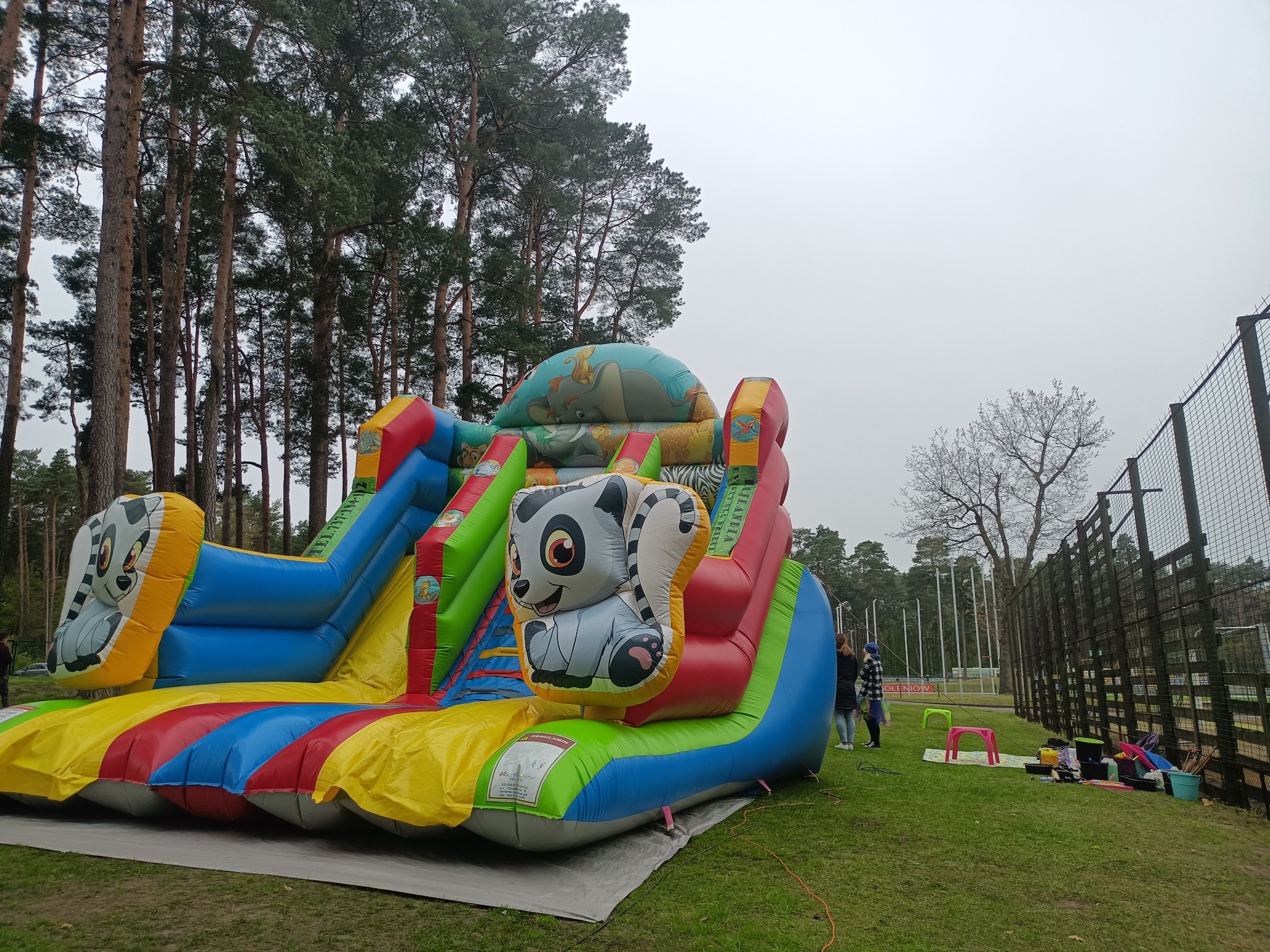 Kolorowy dmuchany zamek ze zjeżdżalniami, z motywem zwierząt, ustawiony na trawie w otoczeniu drzew. Widoczne osoby i elementy infrastruktury.