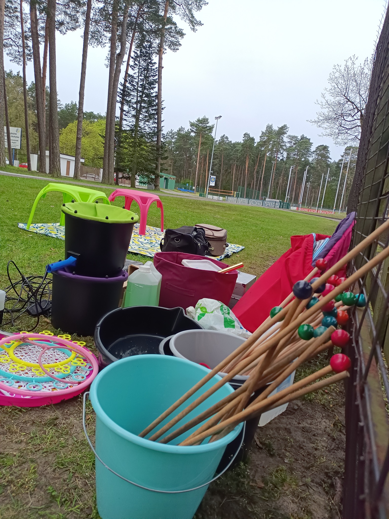 Akcesoria eventowe rozłożone na trawie: dmuchawka do baniek, kije z kolorowymi kulkami, wiaderka, stoliki dziecięce. Przygotowania do imprezy plenerowej.