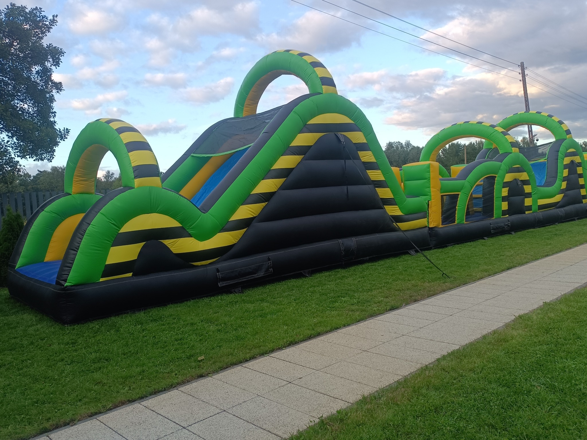 Długi dmuchany tor przeszkód w kolorach czerni, zieleni i żółci, z elementami do wspinaczki i zjeżdżania, ustawiony na trawie obok chodnika.
