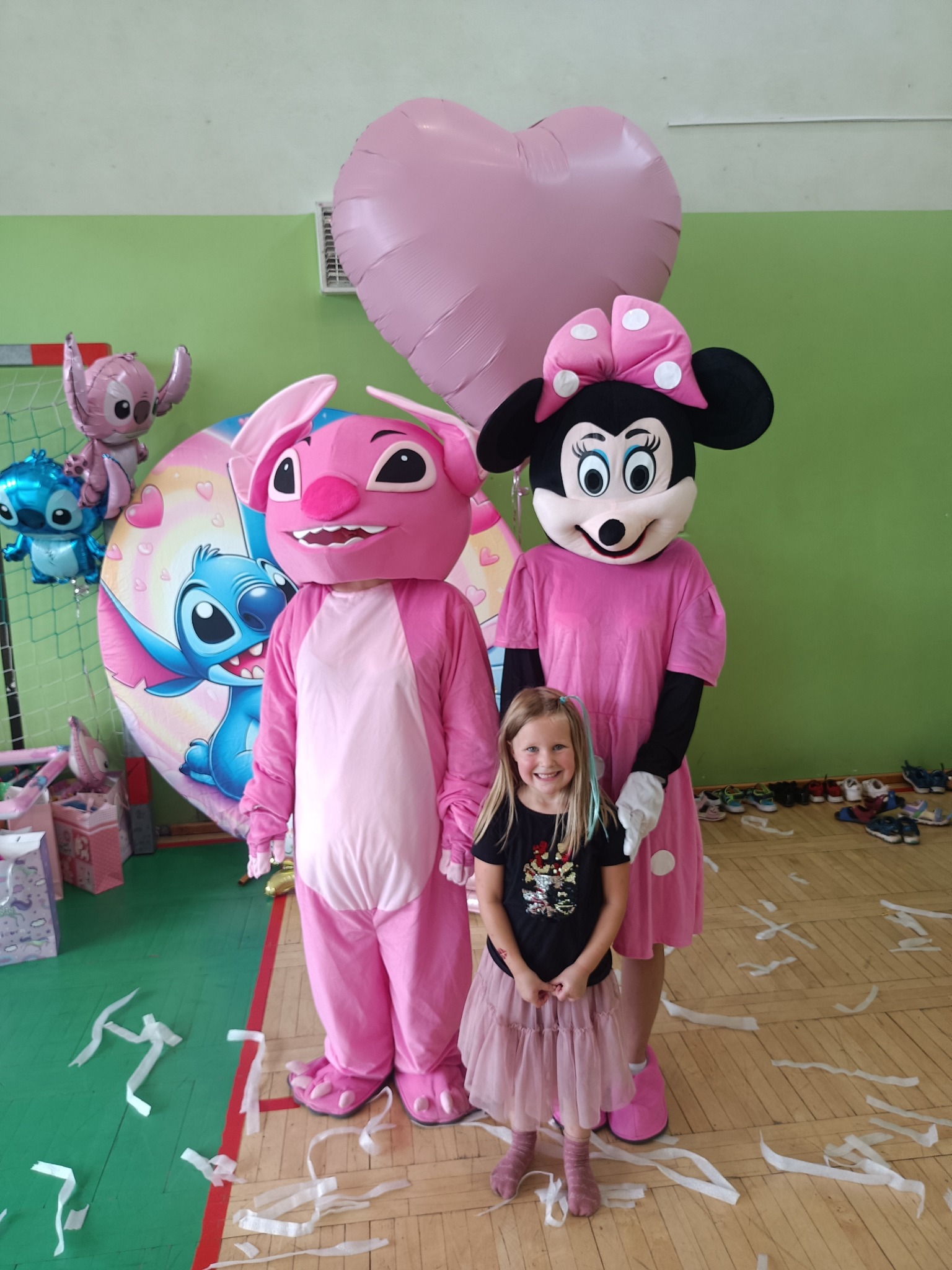 Uśmiechnięta dziewczynka w różowej tiulowej spódniczce pozuje z animatorkami przebranymi za postacie z bajek Lilo i Stich: Angel i Myszka Minnie. Dekoracje balonowe.