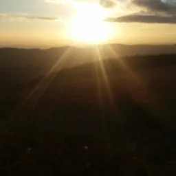 Zachód słońca nad pagórkowatym krajobrazem, promienie słoneczne rozświetlają niebo i wzgórza.