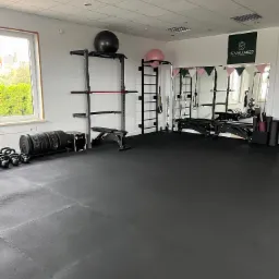 Przestronna siłownia wyposażona w sprzęt do ćwiczeń siłowych, w tym sztangi, kettlebells, ławkę, drabinki i piłki gimnastyczne, z logo 'NE Wellness' widocznym na ścianie.