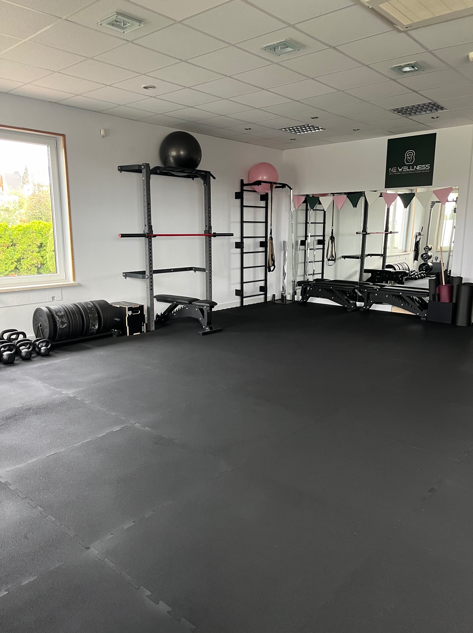 Przestronna siłownia wyposażona w sprzęt do ćwiczeń siłowych, w tym sztangi, kettlebells, ławkę, drabinki i piłki gimnastyczne, z logo 'NE Wellness' widocznym na ścianie.