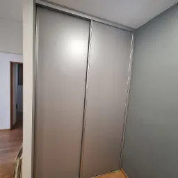 Zamknięta szafa wnękowa z przesuwnymi drzwiami w kolorze szarym, widoczna na tle szarej ściany i drewnianej podłogi. Po lewej stronie szafy oparte o ścianę płyty meblowe.