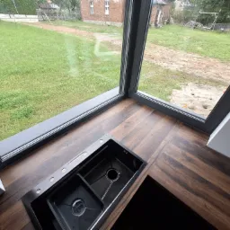 Czarny, dwukomorowy zlew wpuszczony w blat z ciemnego drewna, z widokiem na zielony trawnik za oknem.