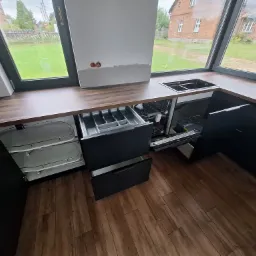 Częściowo zamontowana, nowoczesna kuchnia z czarnymi szafkami, drewnianym blatem i widokiem na ogród przez duże okno. Otwarta szuflada na sztućce i widoczna zmywarka w trakcie montażu.