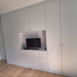 P&P meble Sp.z o.o. - Jasnoszara szafa wnękowa z wbudowanym miejscem na telewizor i szufladami, okrągłe uchwyty, parkiet na podłodze, niebieska ściana w tle.