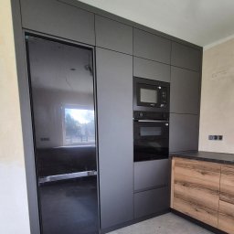 P&P meble Sp.z o.o. - Nowoczesna kuchnia na wymiar w odcieniach szarości z czarnym sprzętem AGD i drewnianymi akcentami. Minimalistyczny design, widoczne niedokończone wykończenie ścian.