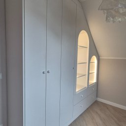 P&P meble Sp.z o.o. - Biała szafa wnękowa z oświetlonymi półkami w kształcie łuków, dopasowana do skosu poddasza, z drewnianą podłogą w jodełkę.