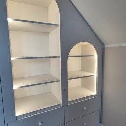 P&P meble Sp.z o.o. - Niebieska szafa wnękowa z oświetleniem LED, wkomponowana w skos dachu, z półkami w kształcie łuków i szufladami z okrągłymi uchwytami.