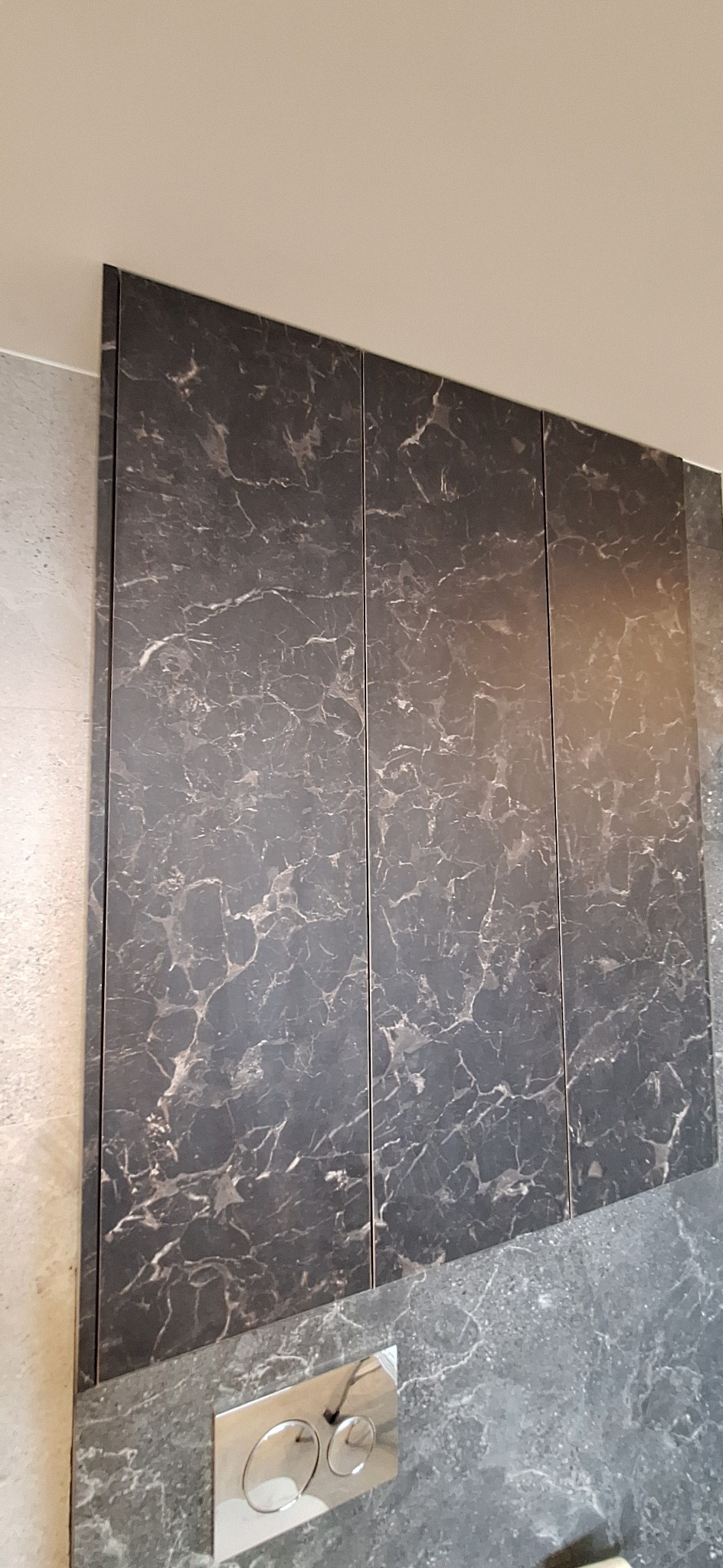 Szafka wisząca z frontami imitującymi marmur w łazience, obok widoczny fragment ściany z szarych płytek i przycisk spłuczki.
