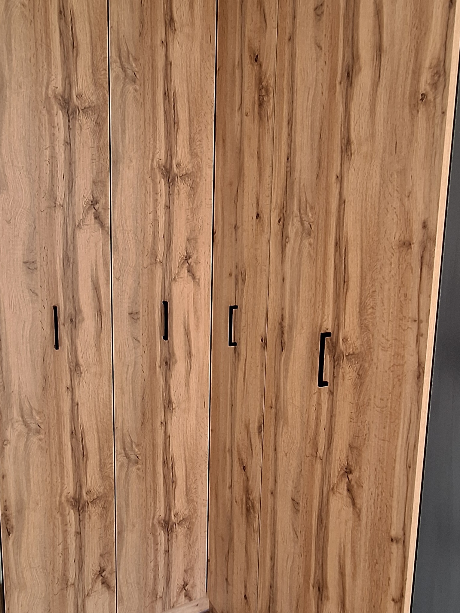 Czterodrzwiowa szafa z frontami w okleinie drewnopodobnej, z minimalistycznymi, pionowymi uchwytami, widoczny fragment bocznej ściany w kolorze grafitowym.