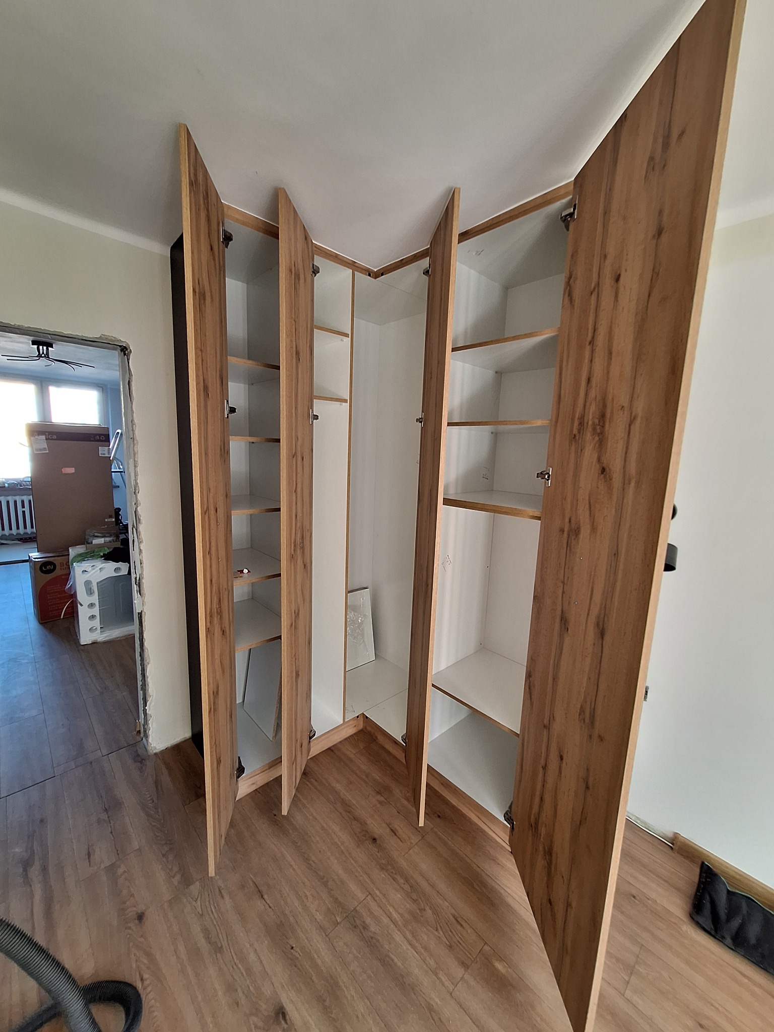 Kątowa szafa wnękowa z drewnopodobnymi frontami i białymi półkami, widoczna podczas montażu w mieszkaniu w trakcie remontu, z elementami wykończeniowymi i odkurzaczem widocznymi na podłodze.