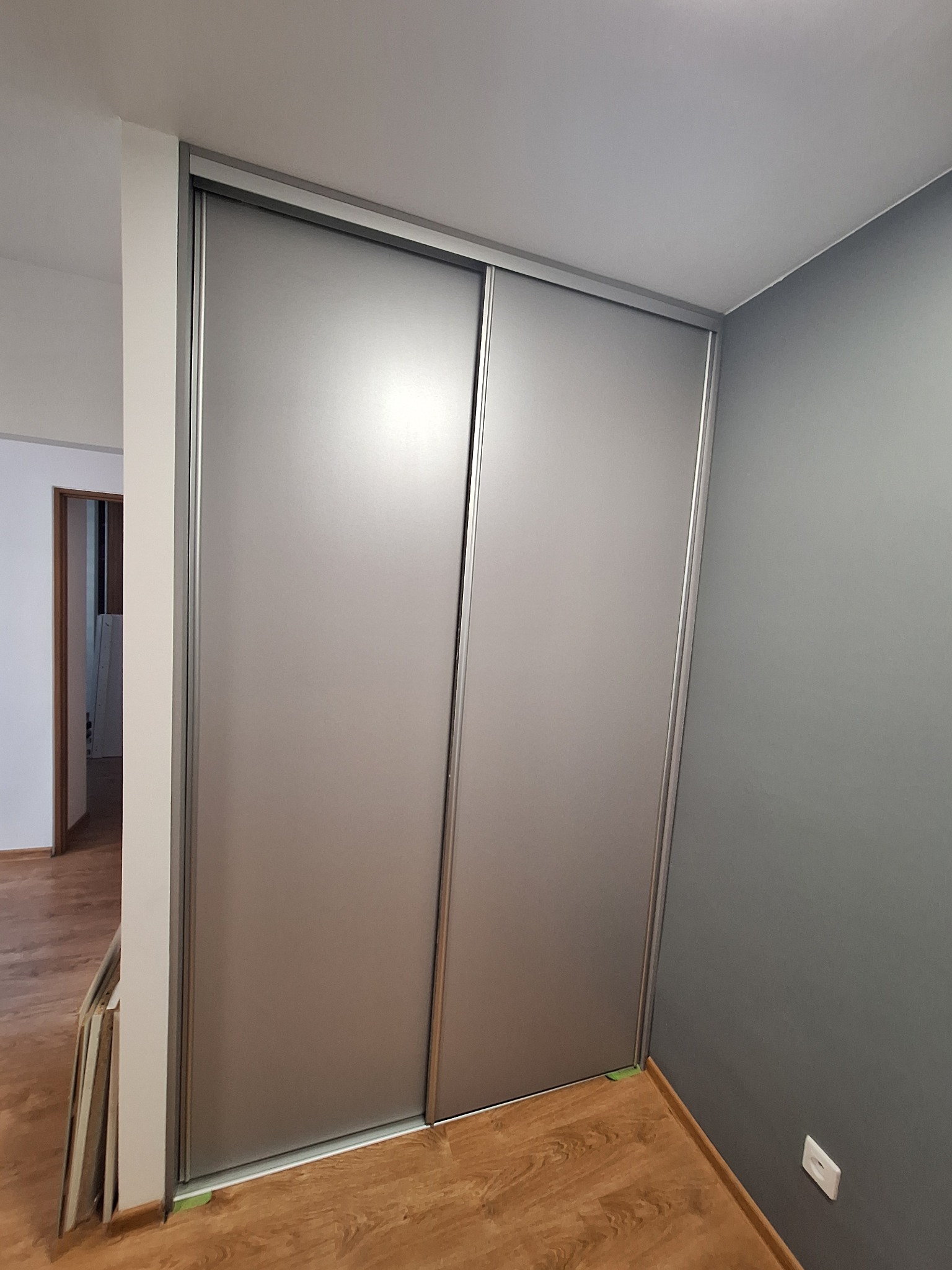 Zamknięta szafa wnękowa z przesuwnymi drzwiami w kolorze szarym, widoczna na tle szarej ściany i drewnianej podłogi. Po lewej stronie szafy oparte o ścianę płyty meblowe.