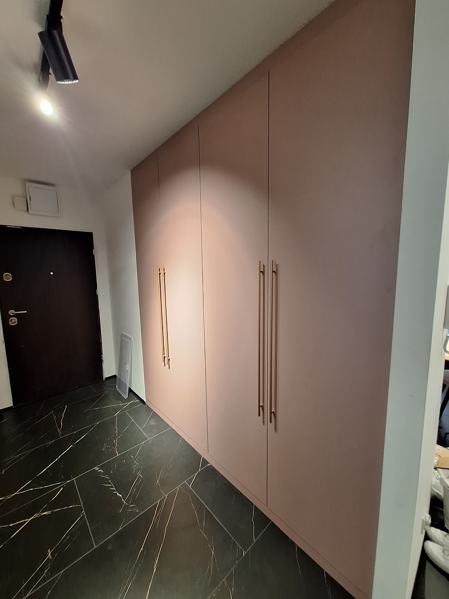 Długa szafa wnękowa z różowymi frontami i złotymi uchwytami, czarna marmurowa podłoga, widok z boku na korytarz z drzwiami wejściowymi.