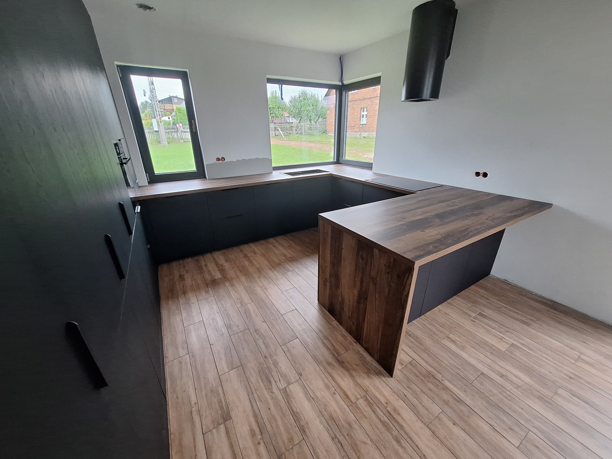 Nowoczesna kuchnia z czarnymi szafkami, drewnianym blatem i drewnopodobną podłogą, z oknami wychodzącymi na ogród i częściowo zamontowanym okapem.