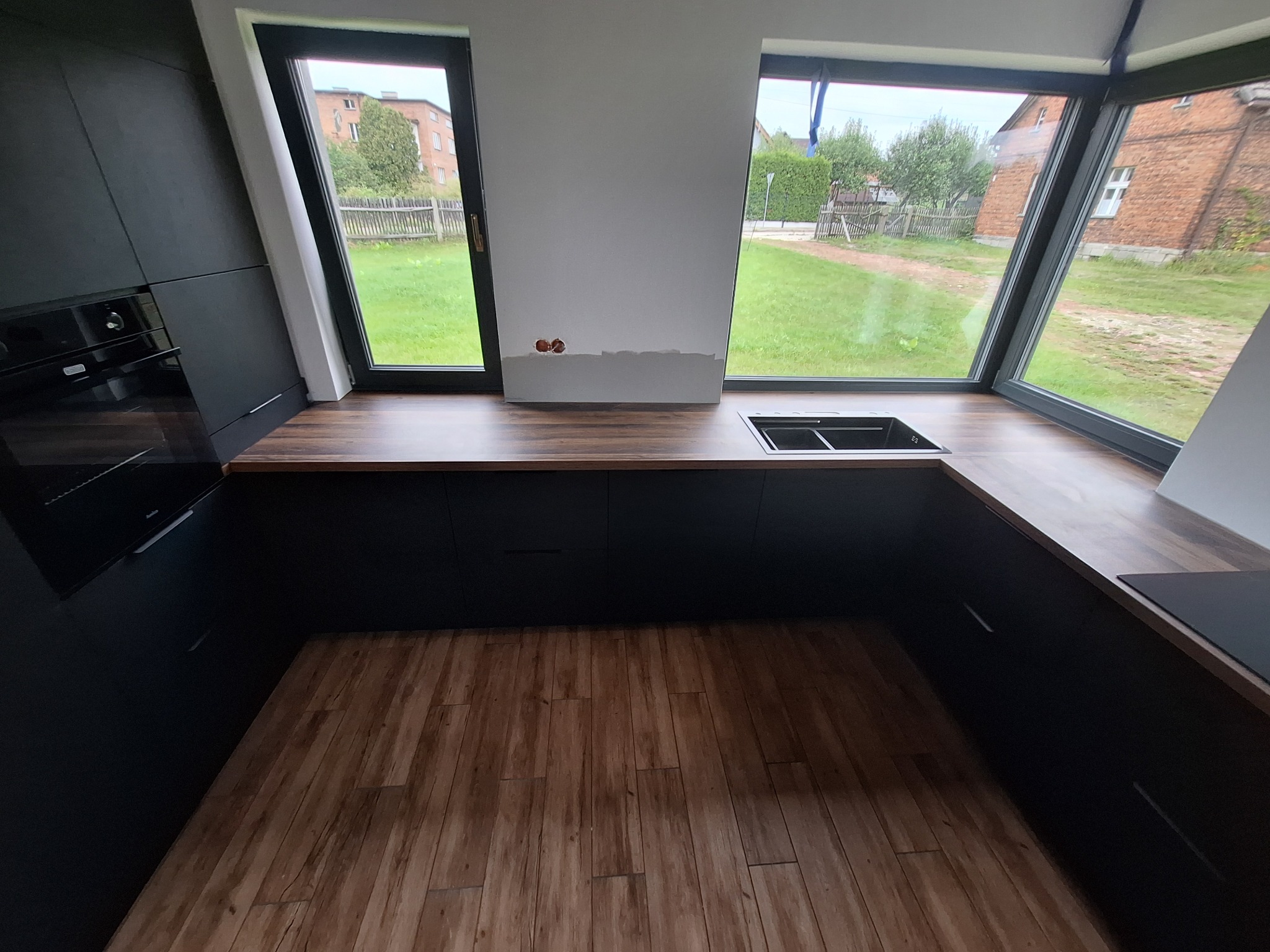 Nowoczesna kuchnia w trakcie montażu, z czarnymi szafkami i drewnianym blatem, widok na ogród przez duże okna, ściana przygotowana pod kafelki.