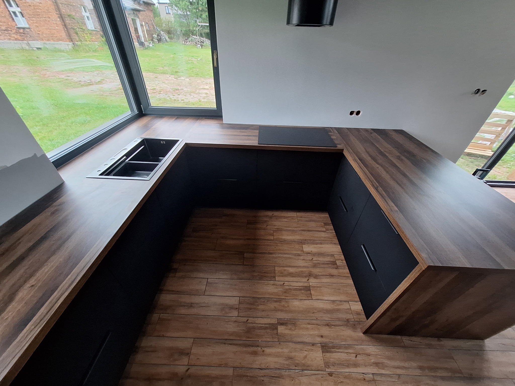 Nowoczesna kuchnia z czarnymi szafkami i drewnianym blatem, zlew wkomponowany w blat pod oknem, widok na zewnątrz, drewniana podłoga, czarny okap, gniazdka elektryczne bez osprzętu.