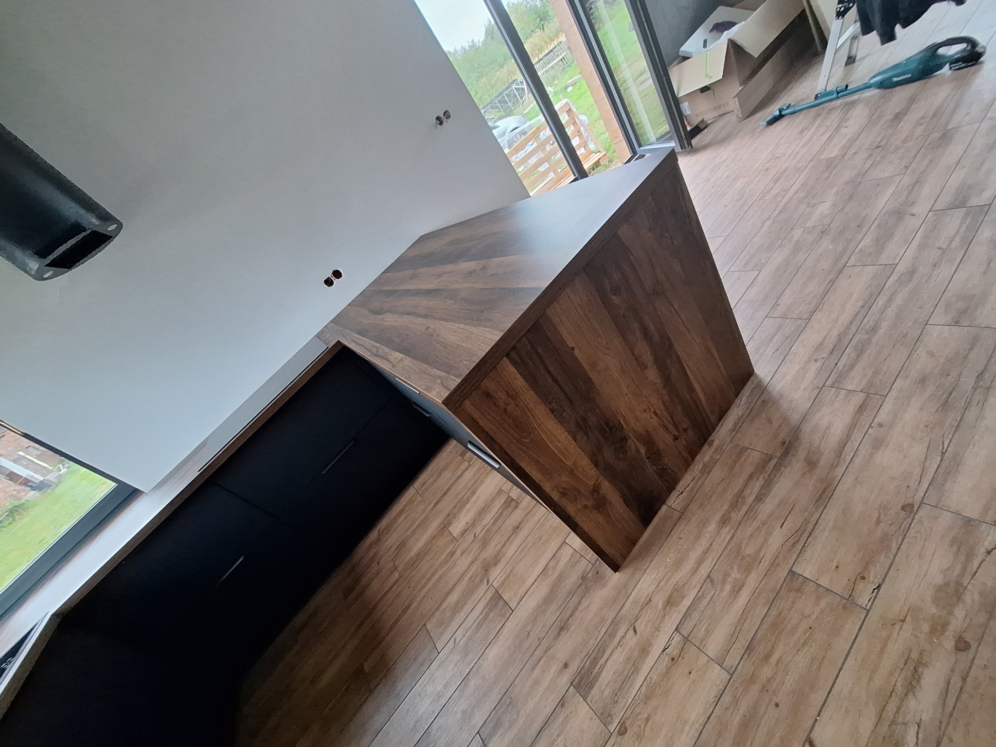 Nowoczesna zabudowa kuchenna z ciemnymi frontami i blatem w odcieniu drewna, widok z góry na fragment pomieszczenia z dużym oknem i elementami wykończeniowymi.