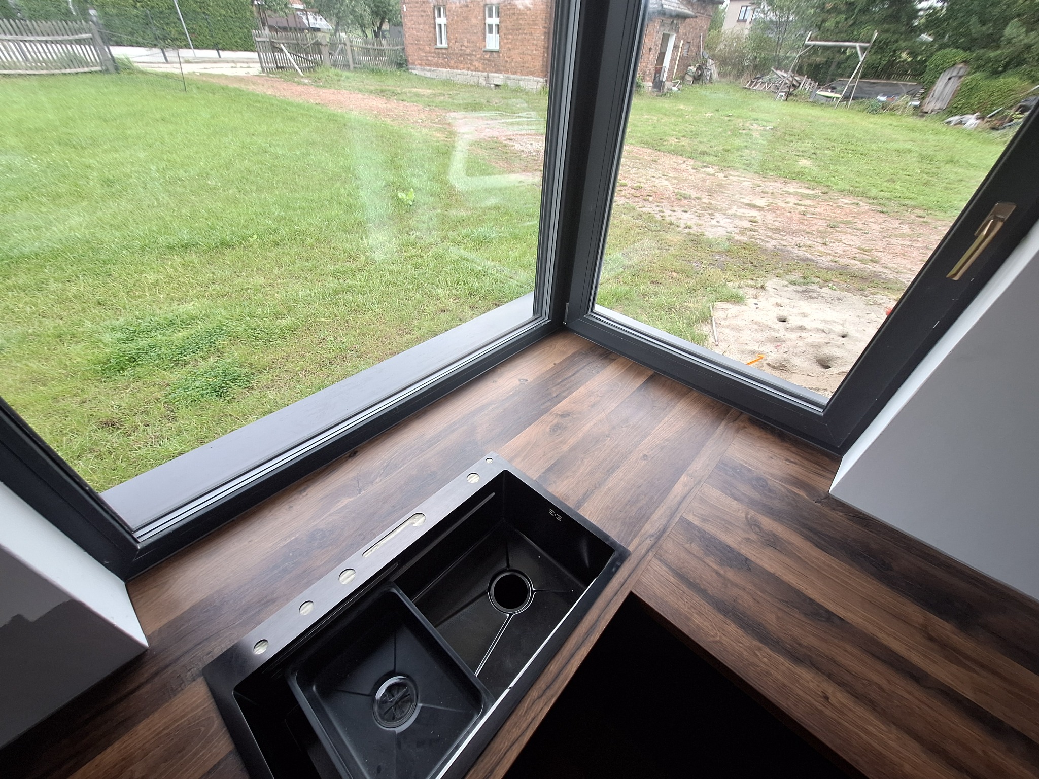Czarny, dwukomorowy zlew wpuszczony w blat z ciemnego drewna, z widokiem na zielony trawnik za oknem.